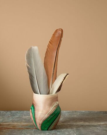 Un jarrón verde y crema con plumas sobre una mesa frente a una pared pintada de color canela.