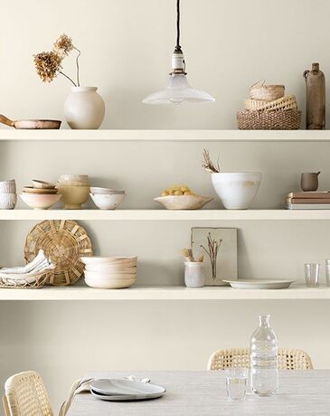 Estanterías de cocina con tazones blancos; mesa y sillas blancas, una pared pintada de blanco