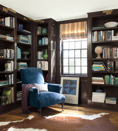 A deep brown library with built in shelves, an animal hide rug, and dark blue velvet chair.