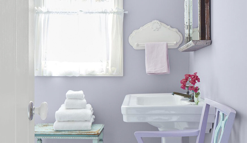 Un baño pequeño y soleado con paredes de color violeta pastel y una ventana con adornos blancos.